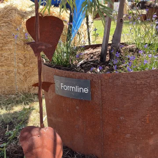 A rusted metal garden stake with heart-shaped blades stands beside a large rusty circular planter. The planter has a dark label reading 'Formline' and is filled with soil, small purple flowers, and tree trunks, with hay bales and grass visible in the background.