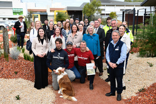 A large group of people posing together outdoors in a landscaped area with gravel, plants, and modern buildings in the background. Several individuals are wearing dark shirts with the text 'Hawthorn Forest', and one person holds a small booklet.